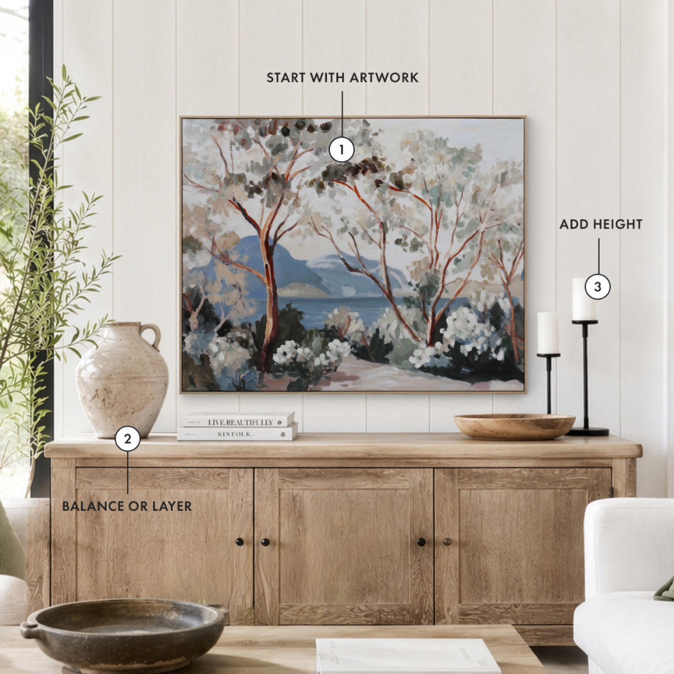 Timber sideboard styled with large landscape artwork, ceramic vase, books, bowl and tall candlesticks, illustrating how scale, varying heights and layered pieces create a beautifully balanced display.