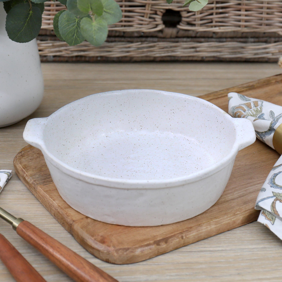 Small round off-white reactive-glaze ceramic baking dish with side handles on a wooden board