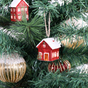 Red ceramic bungalow bauble hanging among Christmas tree branches with gold baubles and another village house