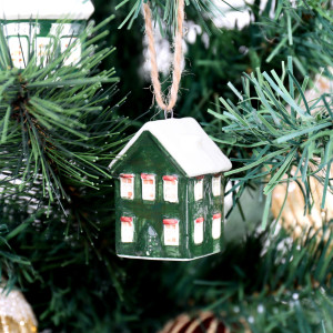 Green ceramic house tree bauble with a snowy white roof and red window accents, hanging among Christmas branches
