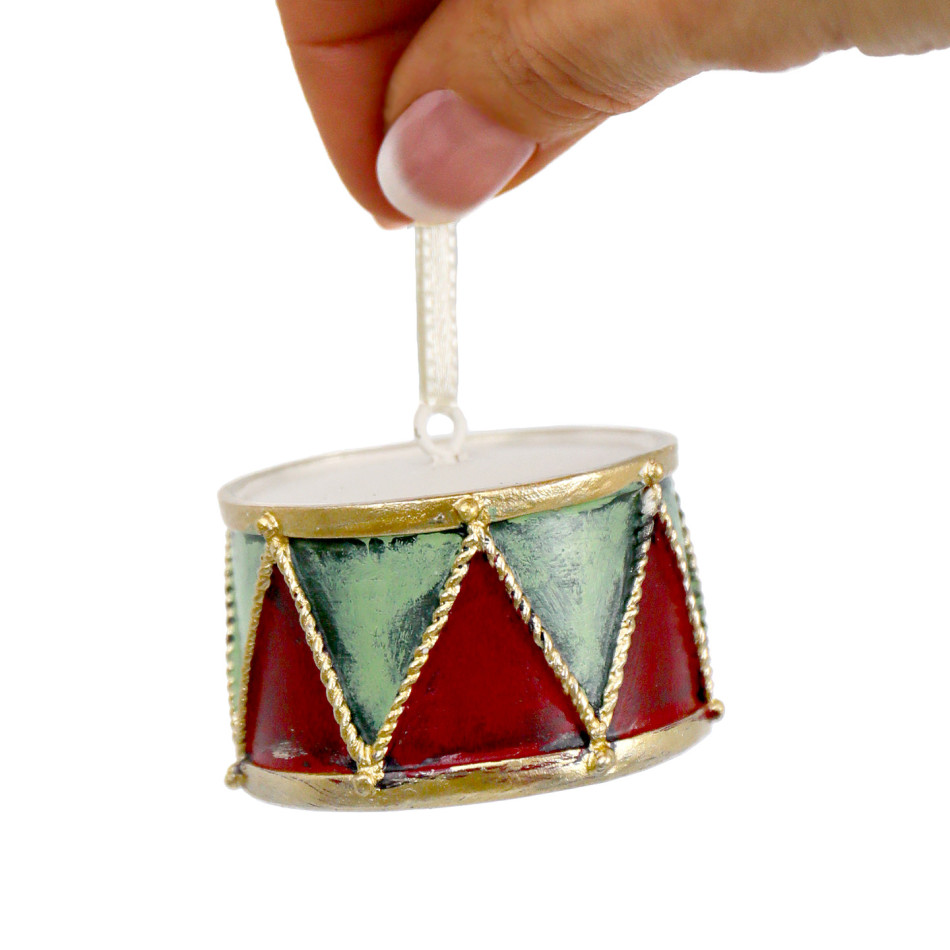 Hand holding a drum Christmas ornament by its pale ribbon, showing triangular panels and raised gold trim