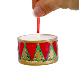 Hand holding the red ribbon of a drum Christmas ornament, showing its pale top and raised gold motifs