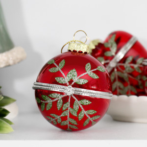 Rosalie round red glass bauble with glittered green leaves and a silver-coloured band on a white surface