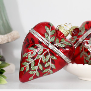 Red glass heart bauble with sparkling green leaves and a decorative metal band beside a white scalloped bowl