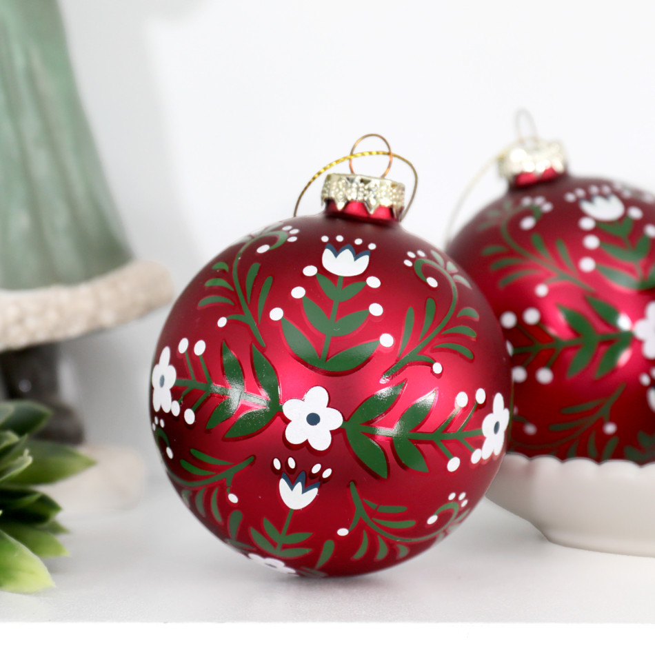 Red glass Christmas baubles with white flowers and green folk-art foliage styled beside a white scalloped bowl