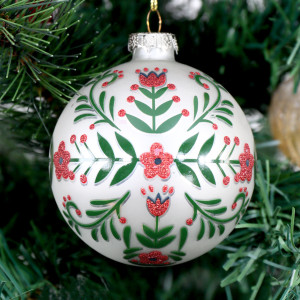 Close-up of white glass Christmas bauble with red glitter flowers and green foliage hanging on a tree