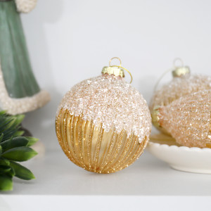 Francesca round gold bauble on a white shelf beside greenery, gold ornaments and a green Santa figure