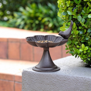 Loville cast iron bird feeder holding seed on the corner of a grey garden wall beside leafy greenery
