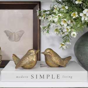 Set of two gold wren figurines displayed on a book beside white flowers and framed butterfly artwork