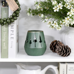 Green forest tealight holder displayed unlit on a white shelf beside pine cones and a vase of white flowers