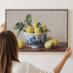 Person positioning a framed lemon still life with a blue-and-white bowl against a white wall