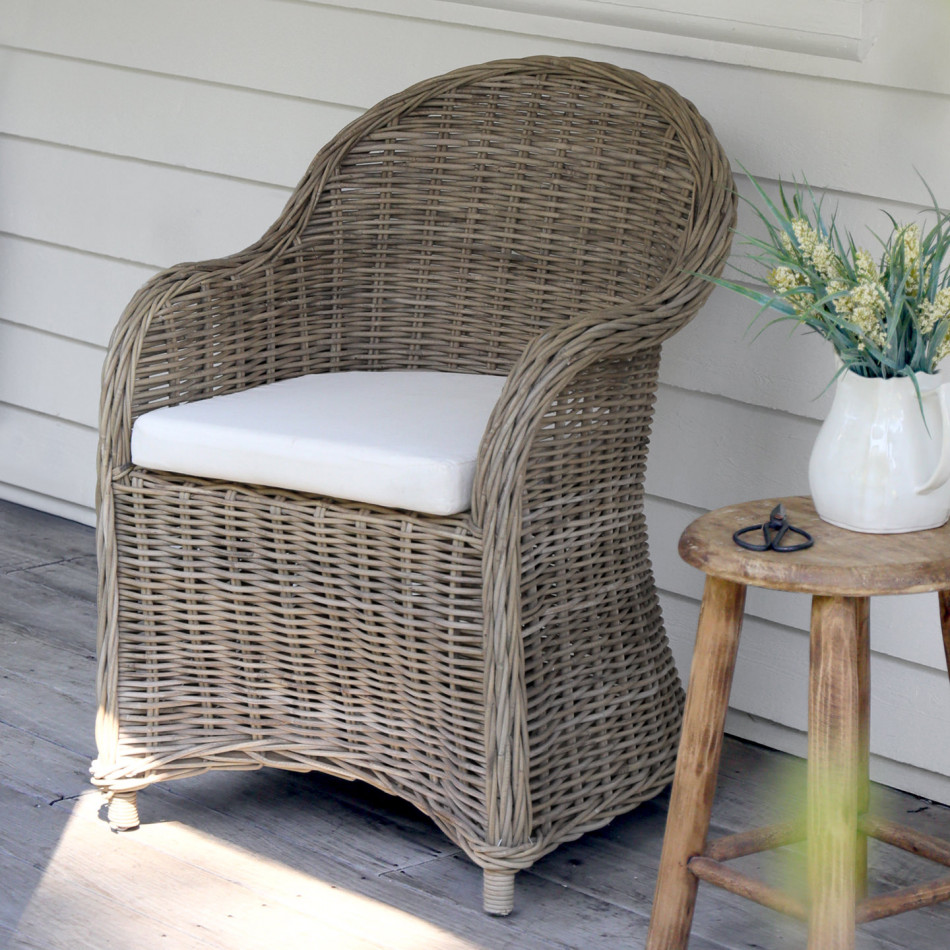 Antique-grey rattan armchair with curved woven back and cream seat cushion on a covered verandah