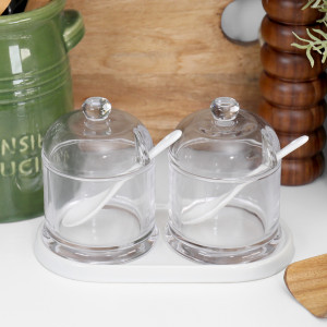 Set of two clear glass condiment jars with white ceramic spoons and base on a kitchen bench