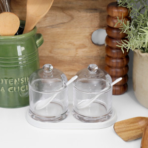 Front view of two glass condiment jars with domed lids, white ceramic spoons and matching base