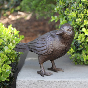 Dark brown cast iron crow standing on a grey garden wall between green shrubs