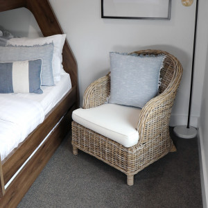 Antique grey rattan armchair with cream cushions positioned beside a wooden bed