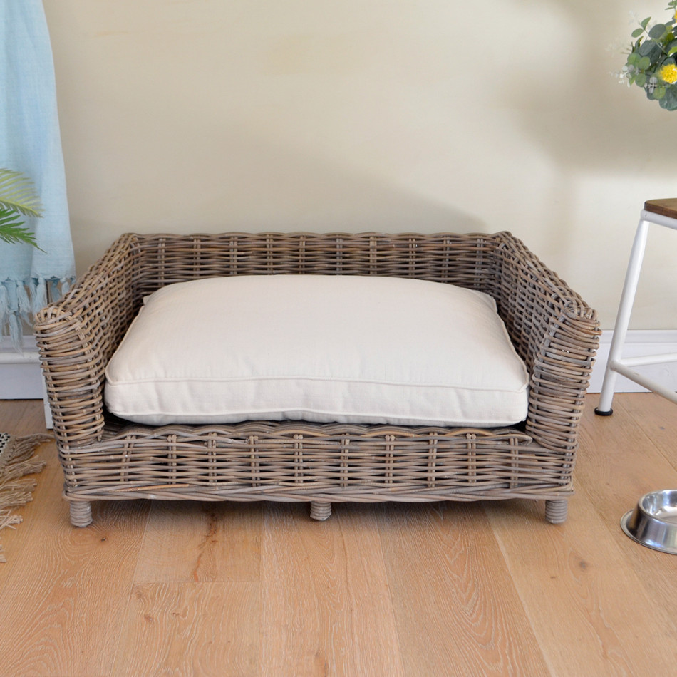 Large antique-grey rattan dog bed with thick natural cream cushion on a wooden floor