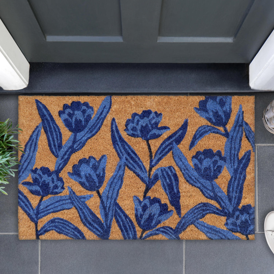 Blue tulip coir doormat on grey tiles beneath a dark front door, with greenery beside it