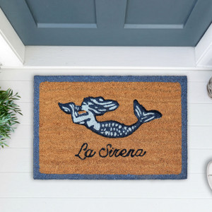 La Sirena mermaid doormat on white floorboards beside greenery, below a blue-grey door