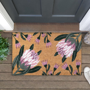 Pink protea doormat at a dark door, styled with greenery, a woven basket and a white shoe on timber boards