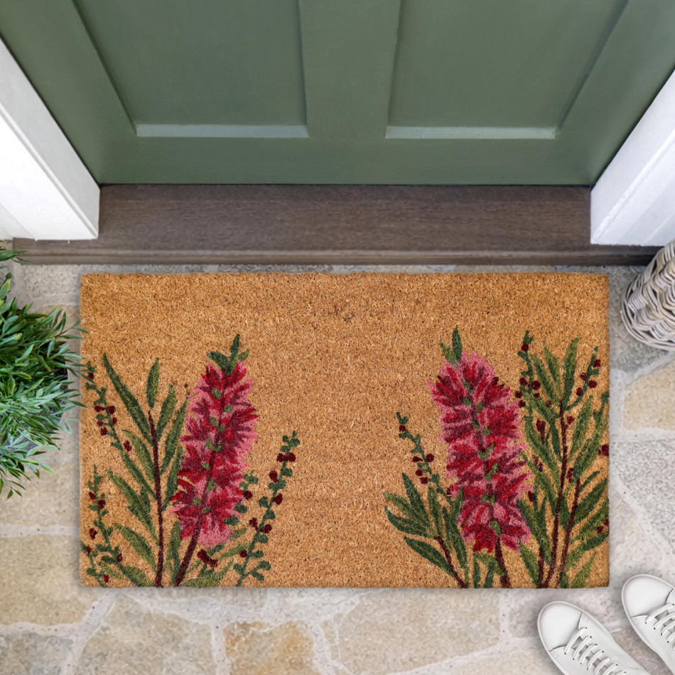 Red bottlebrush door mat on stone paving at a green front door, with foliage and white shoes