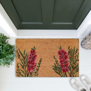Red bottlebrush door mat on white boards at a green door, styled with a leafy plant and woven basket