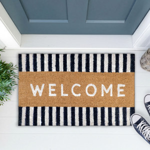Navy-and-white striped welcome doormat at a blue-grey door with greenery, a woven basket and navy shoes