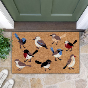 Bird-patterned coir doormat at a green door, styled with greenery, a woven basket and white shoes on stone paving