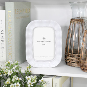 White Sabina ruffle 4x6 photo frame styled with books, white flowers and woven glass jugs on a shelf.