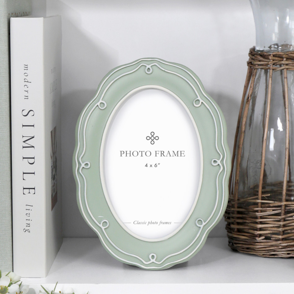Aurelia soft green oval photo frame with raised white detailing styled on a white shelf beside books and a wicker-covered glass vessel.
