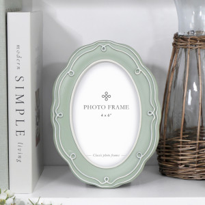 Aurelia soft green oval photo frame with raised white detailing styled on a white shelf beside books and a wicker-covered glass vessel.