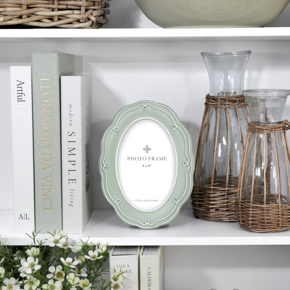Aurelia soft green oval 4x6 photo frame styled on a white bookcase with pale books, wicker-covered glass vessels and white flowers.