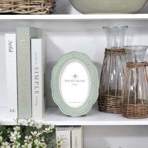Aurelia soft green oval 4x6 photo frame styled on a white bookcase with pale books, wicker-covered glass vessels and white flowers.