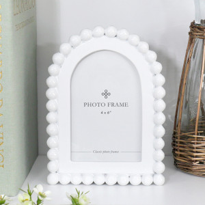 Snowdon beaded white 4x6 photo frame with an arched opening styled on a white shelf beside a pale green book and wicker-wrapped glass jug.