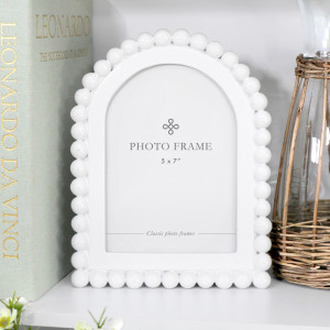 Snowdon beaded white 5x7 photo frame with an arched opening styled on a white shelf beside a pale green book and wicker-wrapped glass jug.