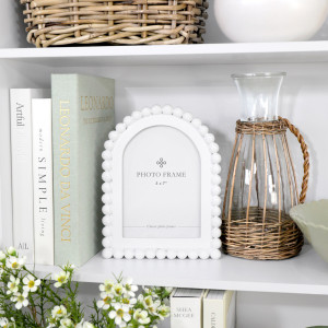 Snowdon beaded white photo frame displayed with the coordinating larger frame, books and wicker-wrapped glass jugs on a white shelf.