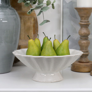 Off-white fluted pedestal fruit bowl filled with green pears in a neutral interior setting.