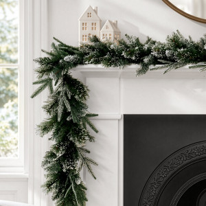 Santa Lucia frosted pine garland draped over the corner of a white mantelpiece beside decorative white houses.