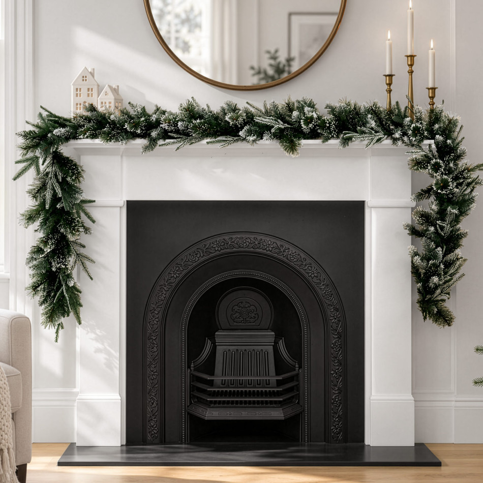 Frosted green pine Christmas garland across a white mantelpiece beneath a round gold mirror, with white houses and gold candlesticks.