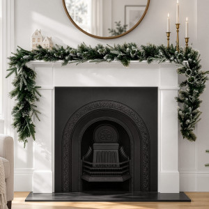 Frosted green pine Christmas garland across a white mantelpiece beneath a round gold mirror, with white houses and gold candlesticks.