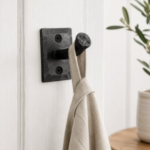 Black iron nail wall hook holding a linen towel on white panelling beside leafy greenery