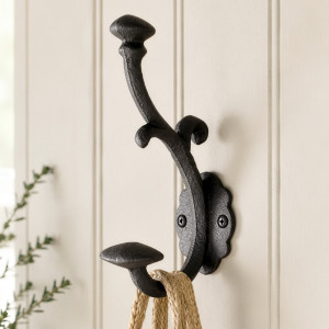 Black cast iron double wall hook with ornate curves mounted on white panelling and holding a woven bag