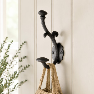 Ornate black double wall hook mounted on white panelling beside leafy greenery