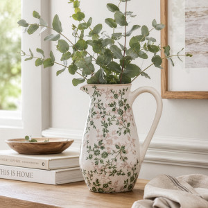 Evergreen floral jug filled with eucalyptus beside books and a folded striped cloth on a timber console