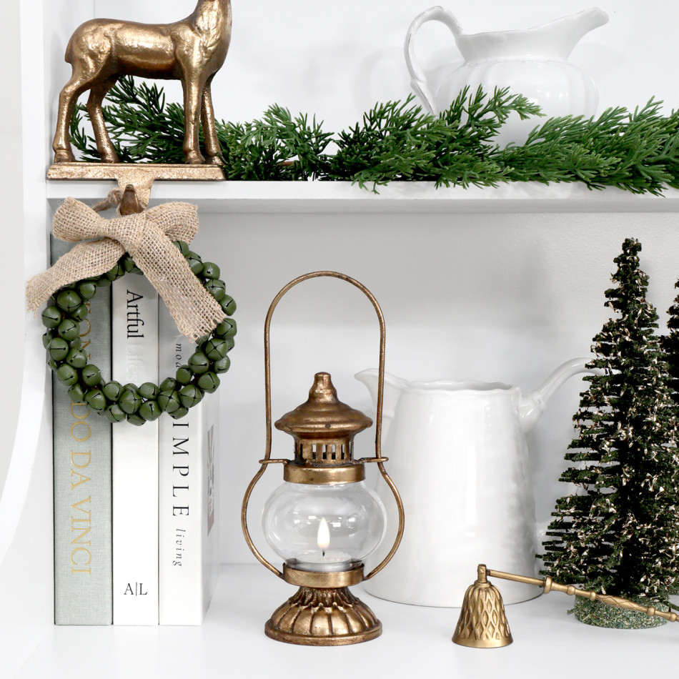 Yuletide gold tealight lantern on a white shelf with a green bell wreath, Christmas trees and a gold candle snuffer.