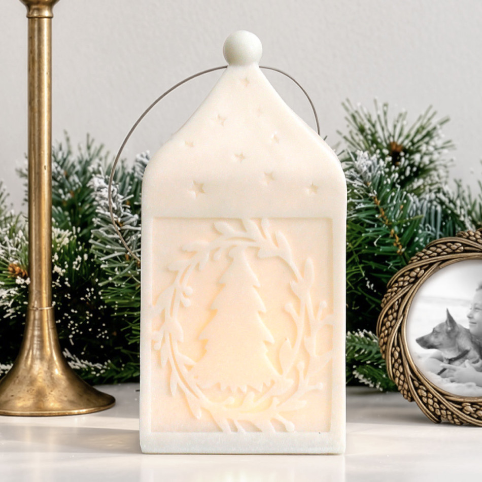 Large white Silent Night lantern illuminated to show its Christmas tree and leafy wreath design, with frosted greenery behind.
