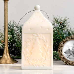 Large white Silent Night lantern illuminated to show its Christmas tree and leafy wreath design, with frosted greenery behind.