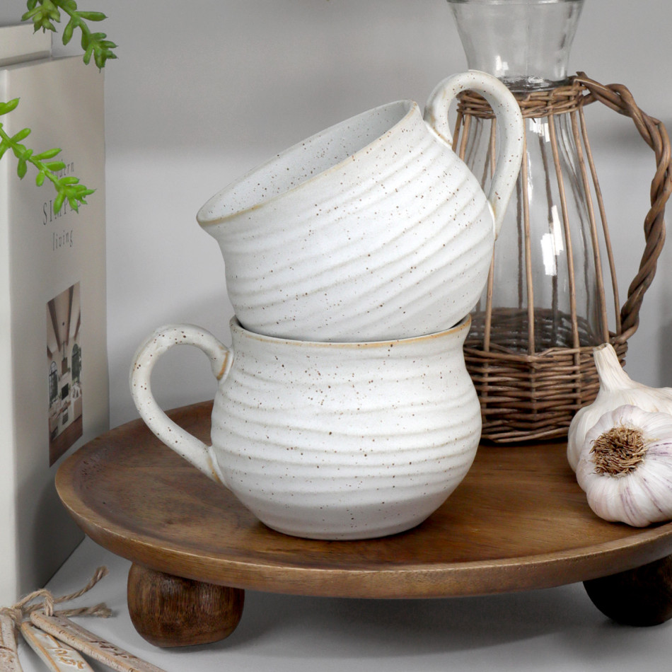 Two speckled off-white ribbed ceramic mugs stacked on a raised wooden board