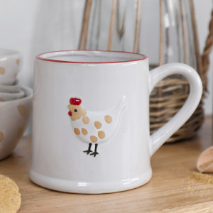 White ceramic mug with red rim and raised hen motif styled on a wooden board beside matching bowls