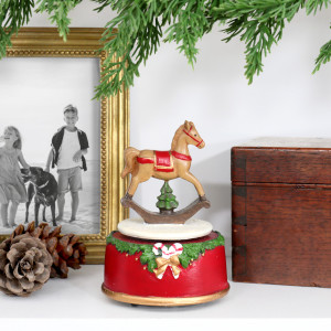 Rocking horse Christmas music box displayed beneath greenery with family photographs, pinecones and a wooden box